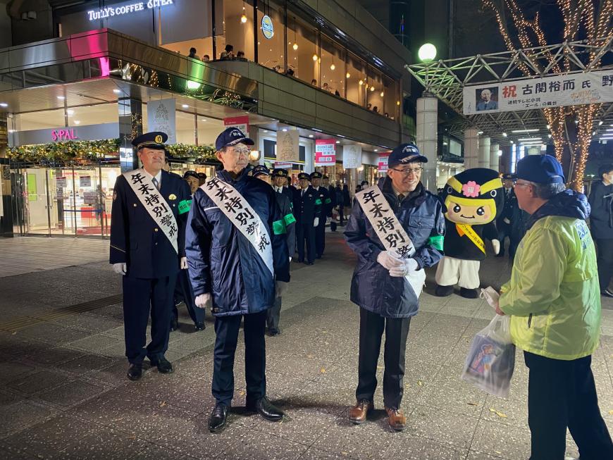 イルミネーションされた夜の街で「年末特別警戒」と書かれた襷をかけた市長と警官が通行している人に話をしている写真