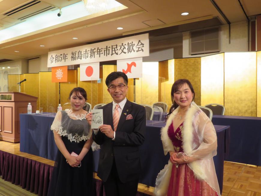 「令和5年福島市新年市民交換会」のボードがある会場のステージ前で、市長の両脇にドレス姿の女性が立っている写真