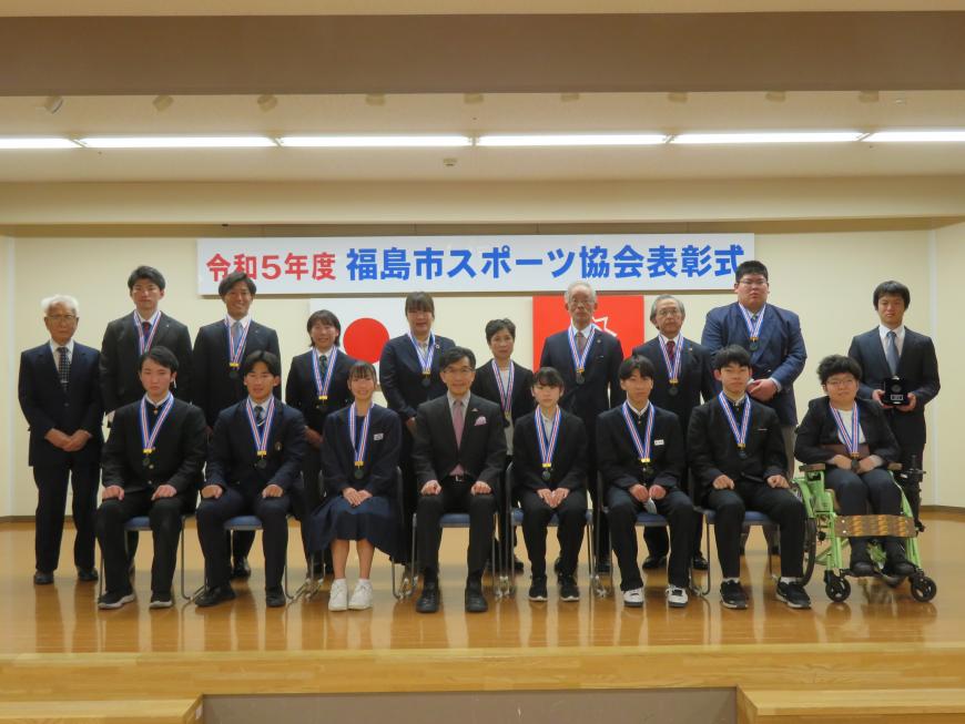 ステージ上で前列に市長を中心にメダルをかけた7名が座り、後方に10名が立っている記念写真