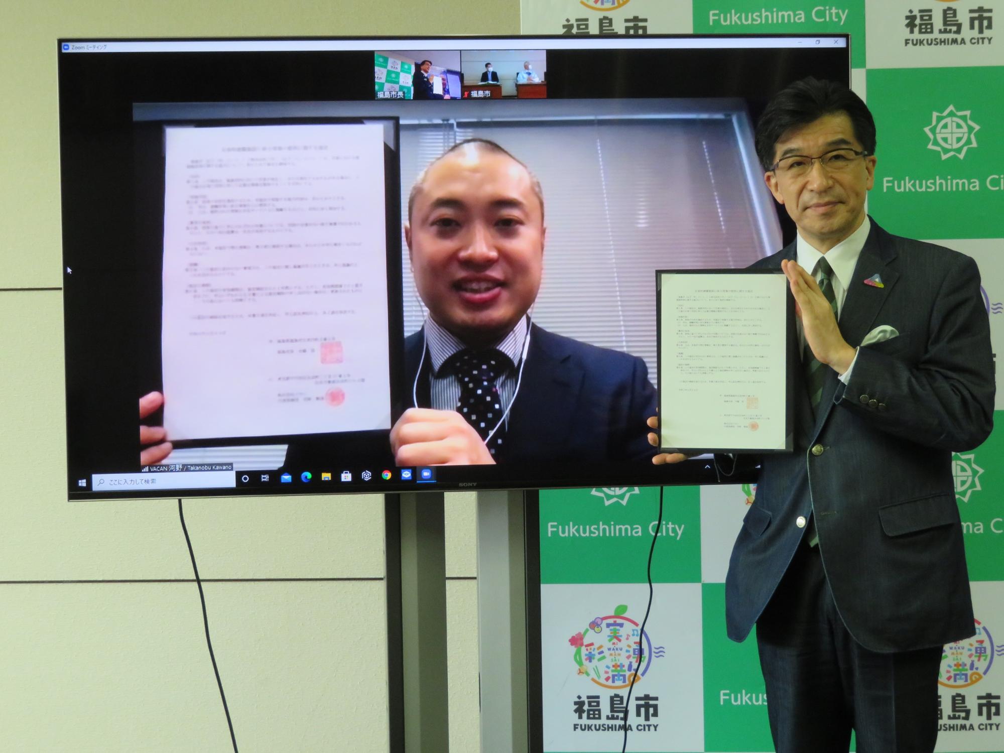 福島市のバックパネル前で、モニター越しに協定書を手に持っている株式会社バカンの関係者と、市長が協定書を手に持っている写真