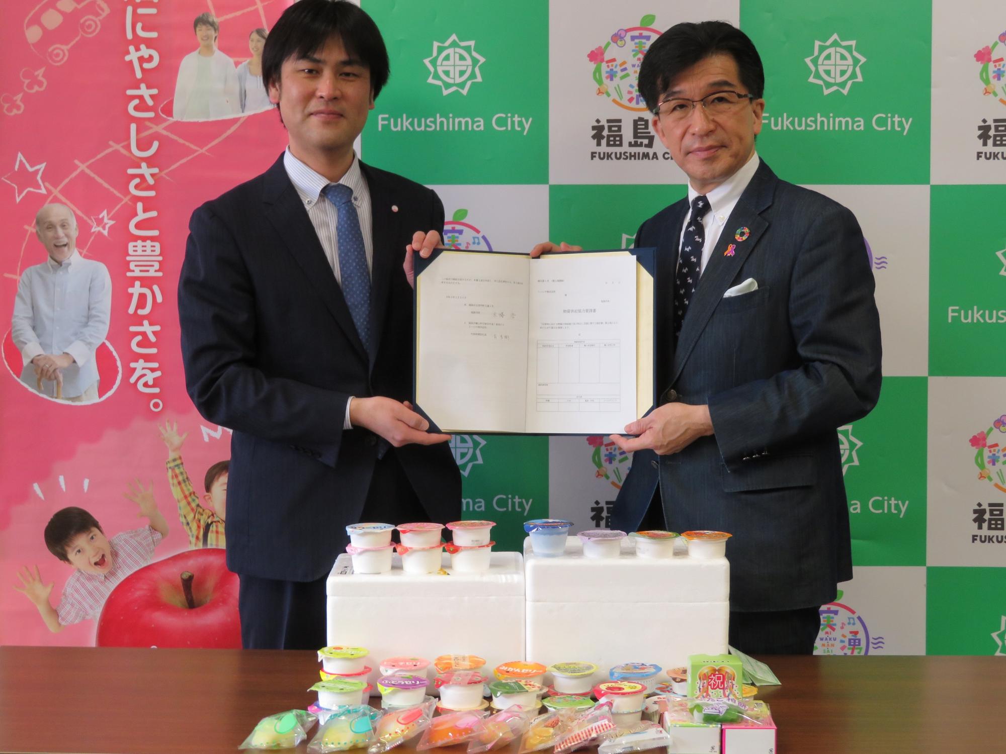 福島市のバックパネル前に、ゼリーなどの食料品が机に並べられており、市長とトーニチ株式会社の関係者が一緒に協定書を手に持っている写真