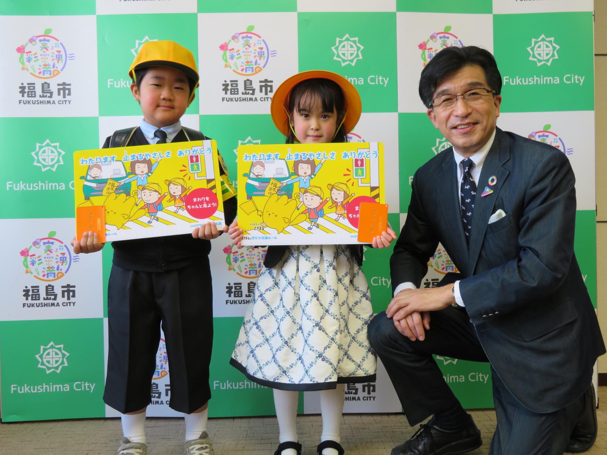 福島市のバックパネル前で、黄色の帽子を被った男の子と女の子が新入学児童交通安全用具を手に持ち、子供達と同じ目線で膝まづく市長の写真