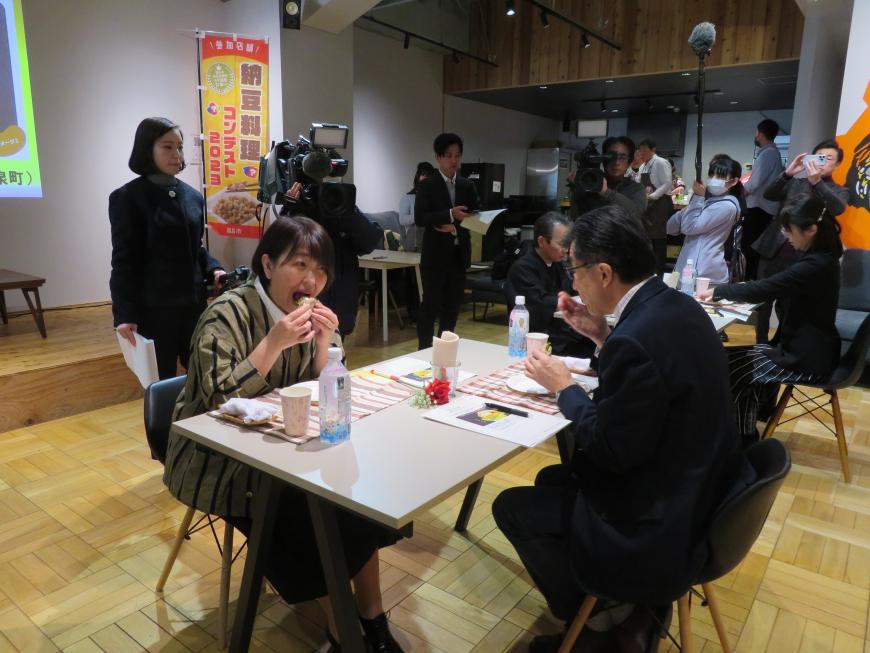 テレビカメラが撮影する中、市長と女性が向かい合って座り、料理を食べている写真