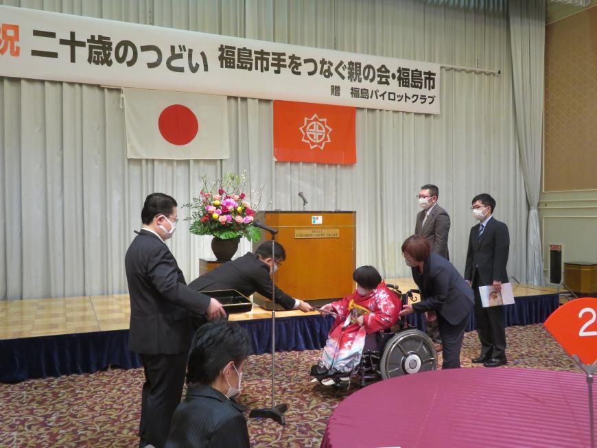参列者が見ている中、市長が車いすの女性に記念品を渡している写真