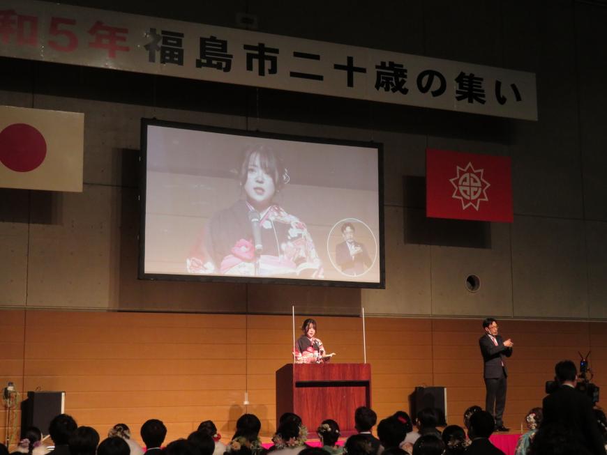 参加者が客席に座っている前で、着物姿の女性が演台で話をしている状況がステージのモニターに映し出されている写真