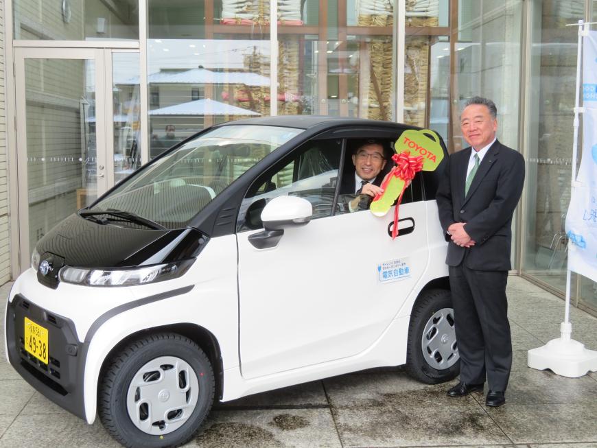 市長が乗車した軽自動車から顔を見せて、鍵のレプリカを持ち、車の隣には男性が立っている写真
