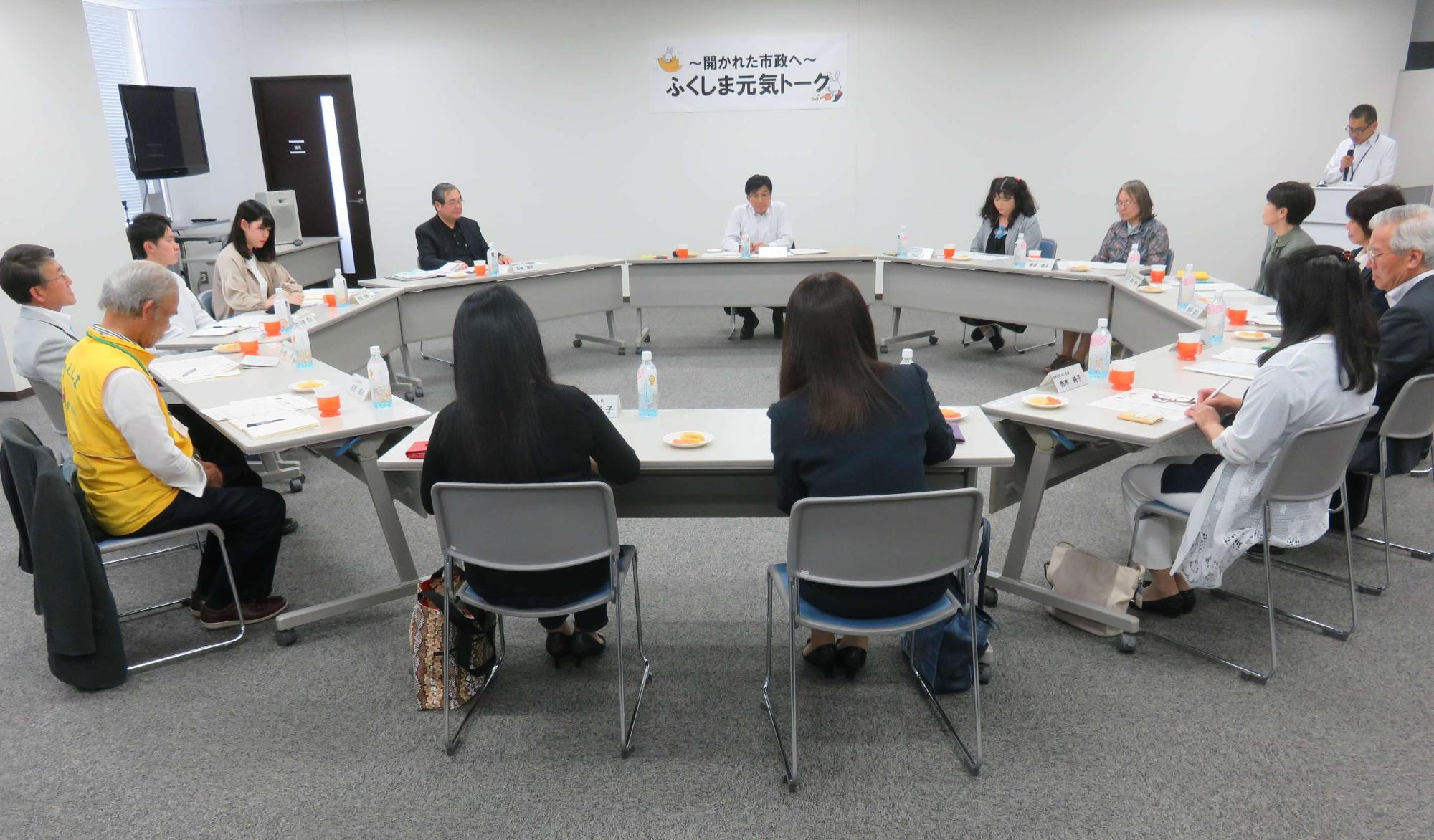 円形に並べられた机に市長と関係者が椅子に座り話し合いが行われている様子の写真