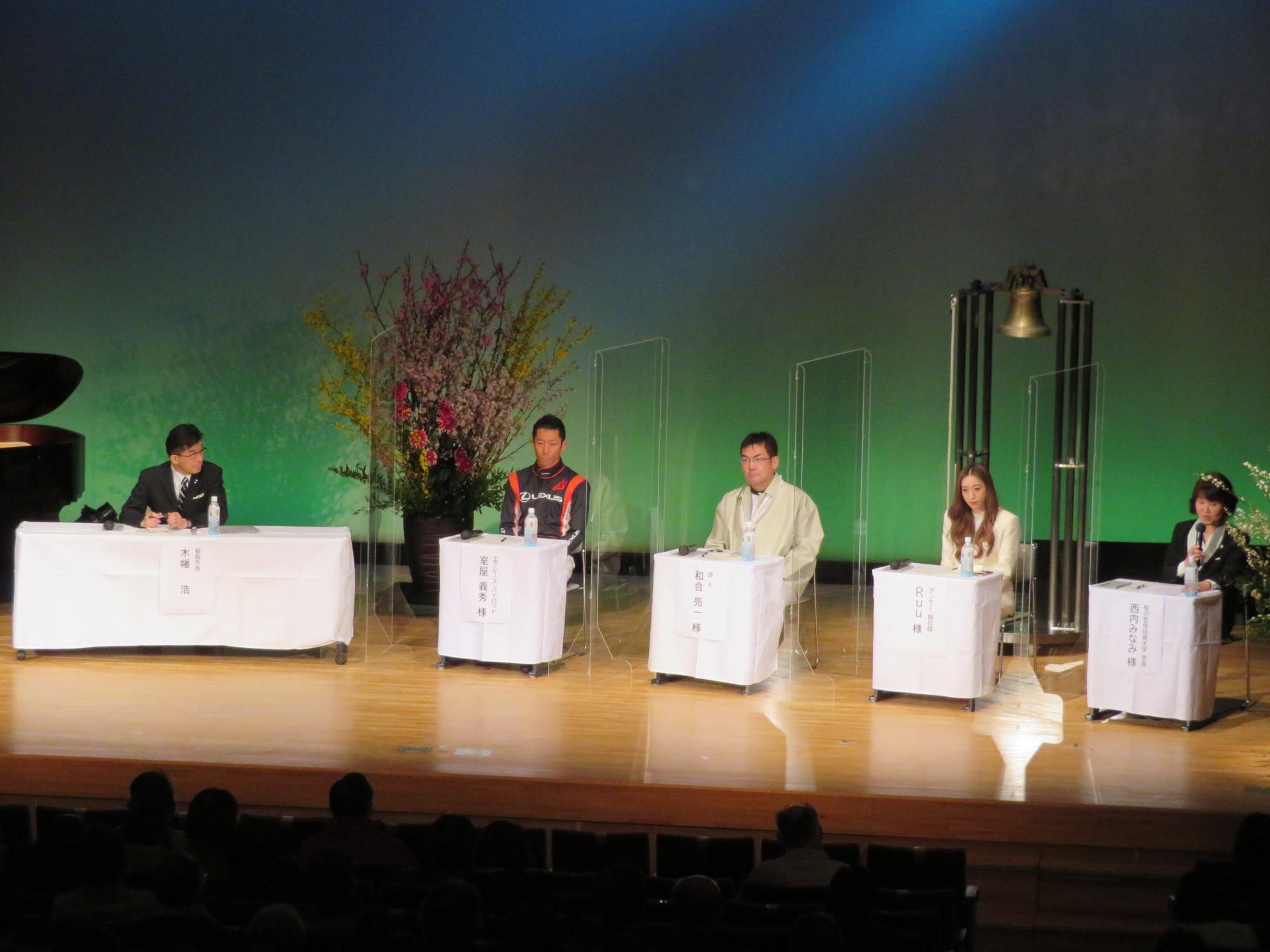 客席に参加者がいる会場で市長と4人のパネリストがステージで話をしている写真