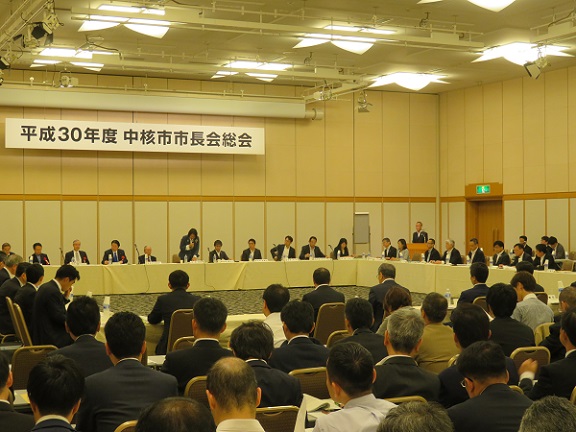 広い会場の一室で、「平成30年度中核市市長会総会」のパネルがあり、コの字型の席で多くの関係者が椅子に座っている写真