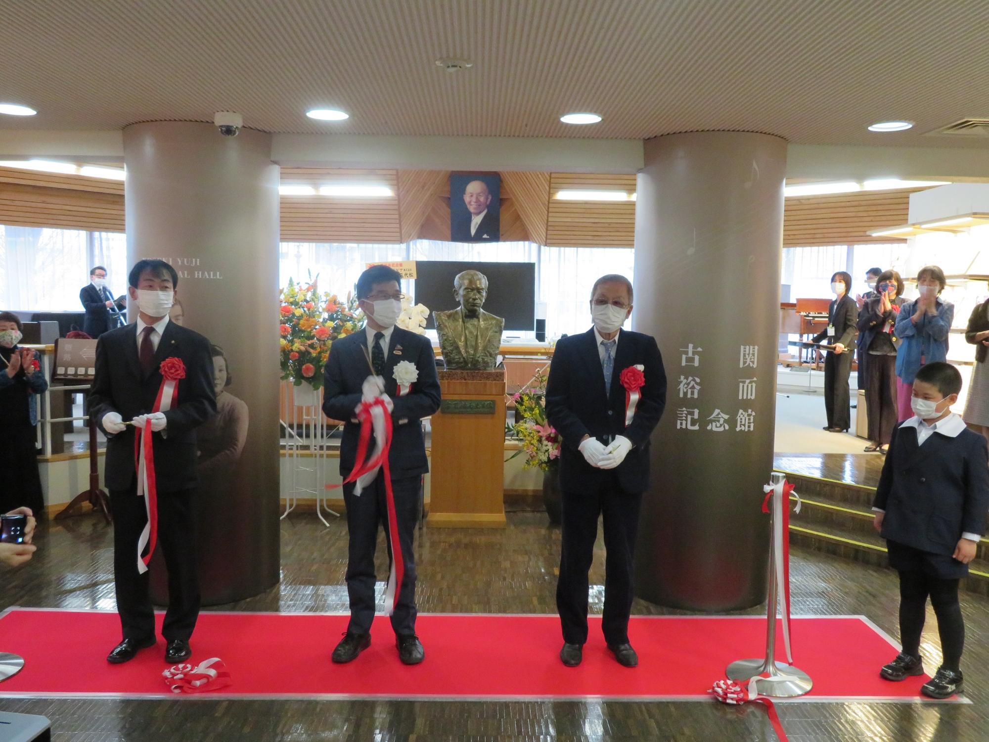 古関裕而さんの写真や銅像があり、柱に古関裕而記念館と書かれた場所で市長を含む男性3人でテープカットをした写真