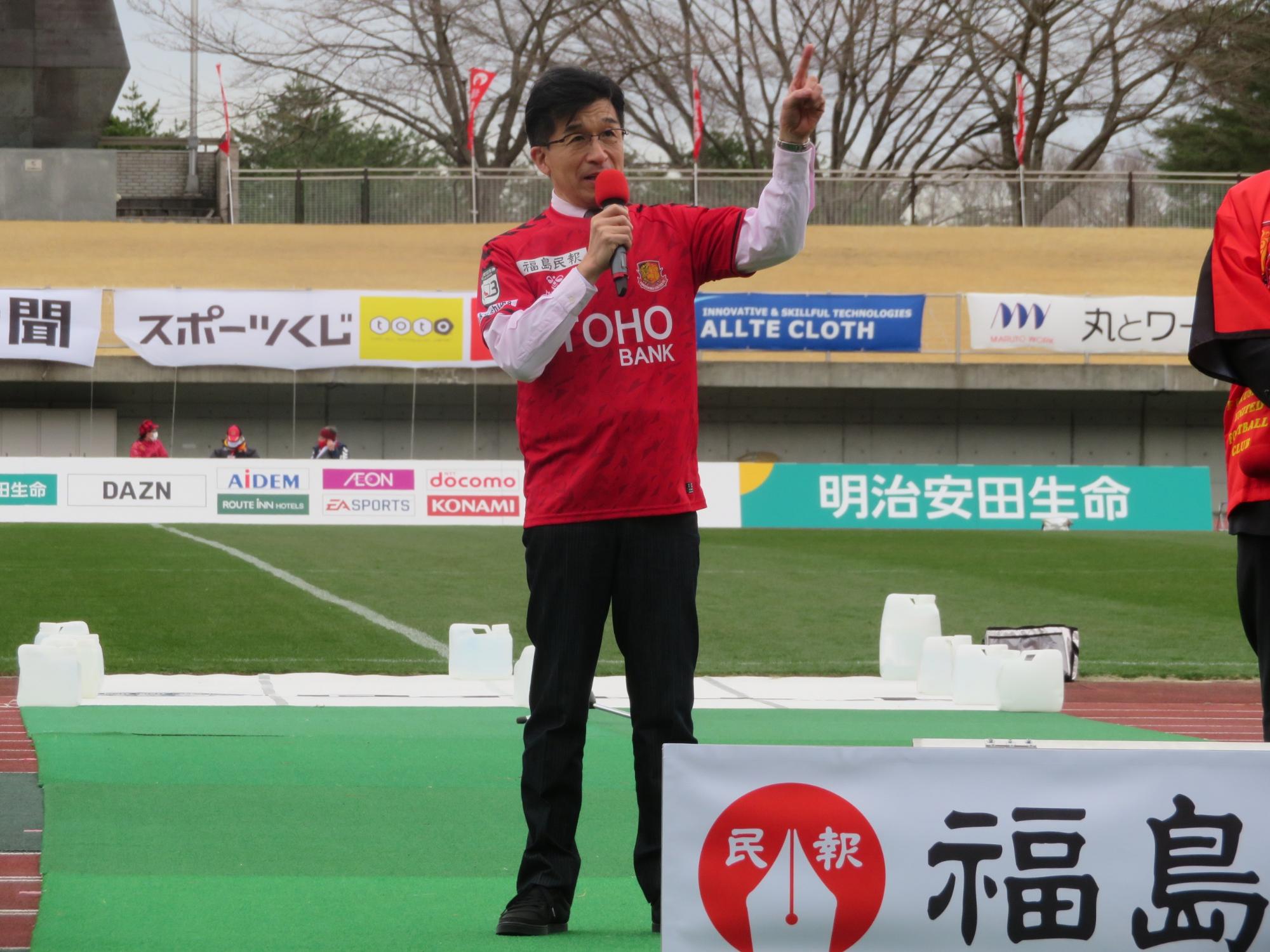 スポンサーの名前が入った横断幕やボードがあるスタジアムで赤のユニフォームを着た市長が左手の人差し指を立てながらマイクで話をしている写真