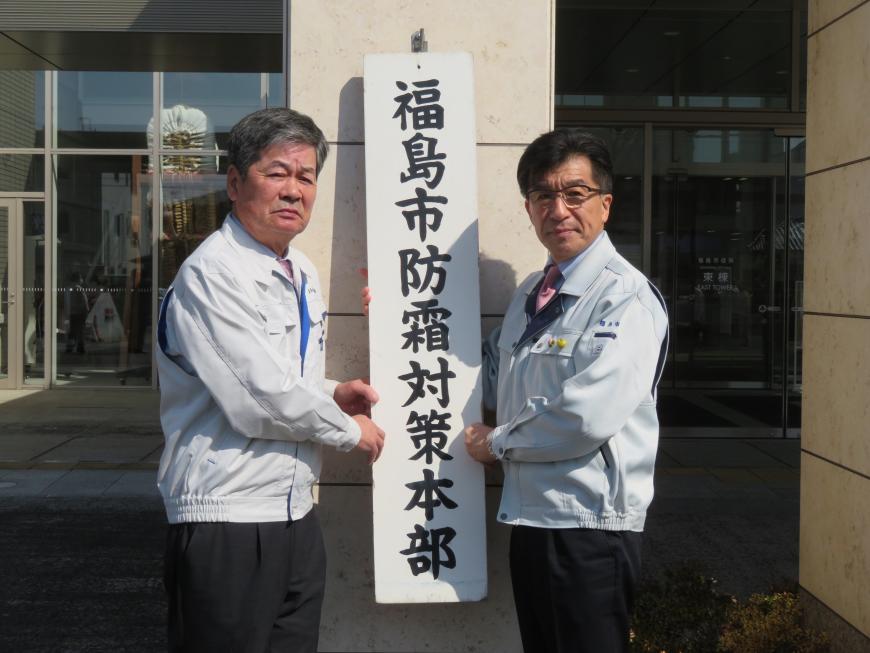 作業用のジャンバーを着た市長と男性で「福島市防霜対策本部」と書かれた看板を屋外の壁にかけている写真