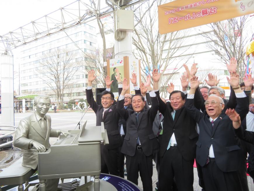 男性がピアノを弾いている銅像の前で、市長を含む関係者の方が万歳をしている写真