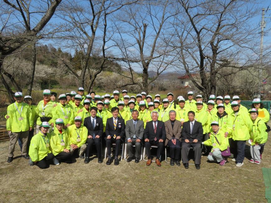 広場の一角でお揃いの帽子と黄色のジャンバーを着た大勢のスタッフの方々と市長を含めた関係者方々が並んだ集合写真