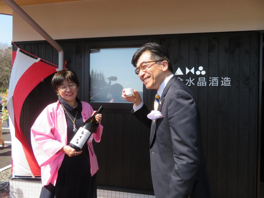 酒蔵の名前が書いてある壁の前で、法被を着て一升瓶を持った女性と、おちょこを持った市長が立っている写真