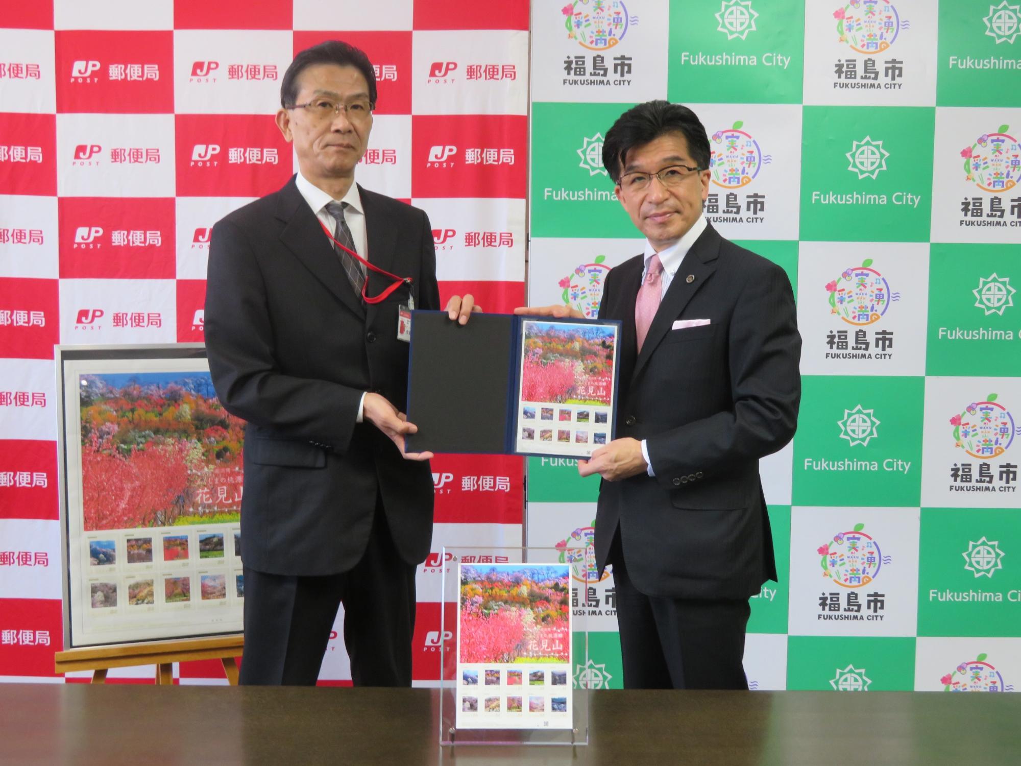 福島市と郵便局のバックパネルの前で、市長と男性で切手が入っているファイルを持って立っている写真