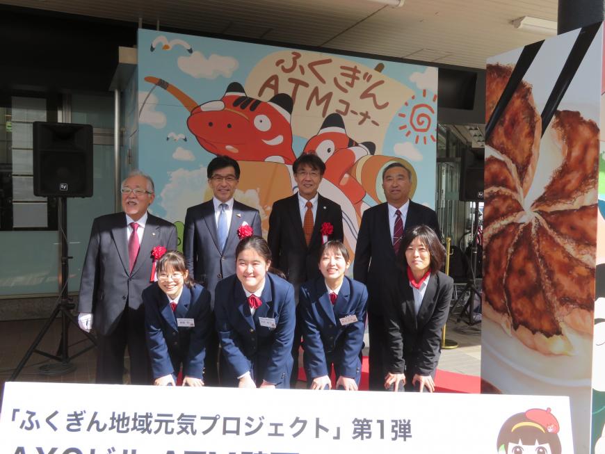 「ふくぎんATMコーナー」の文字が入った壁面アートを背景に、市長を含む男性4名の前に中腰姿勢の女性が4名並んでいる写真