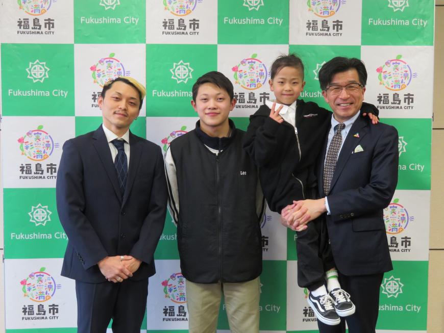 バックパネルの前で、市長が児童を抱きかかえ、男性2名が並んで立っている写真