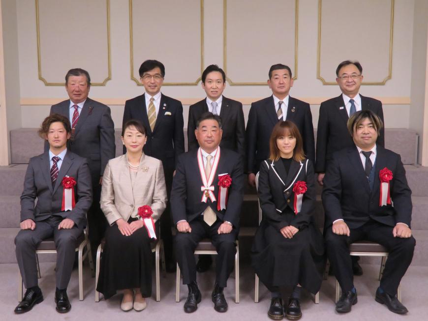 前列にメダルをかけた男性を含めて男性3名、女性2名が座り、後列に市長を含む男性5名が立っている写真