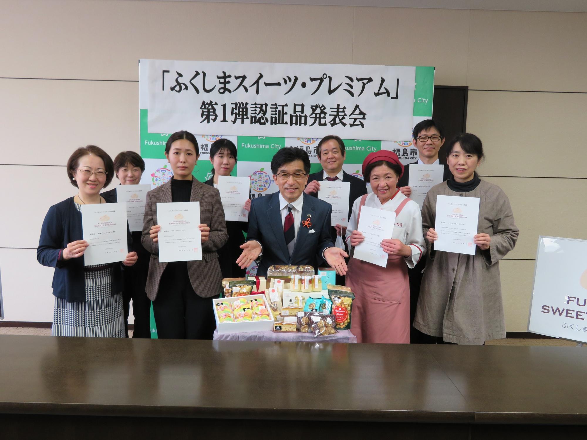 「ふくしまスイーツ・プレミアム」第1弾認証品発表会と書かれた横断幕が貼られたバックパネルの前で、市長の回りに賞状を持った8人が集まり、その前には認証品が置いてある写真