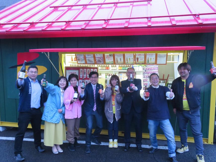 赤い屋根の店先で市長を含む男女8名がそれぞれビール、日本酒、ワインの瓶を持って並んでいる写真