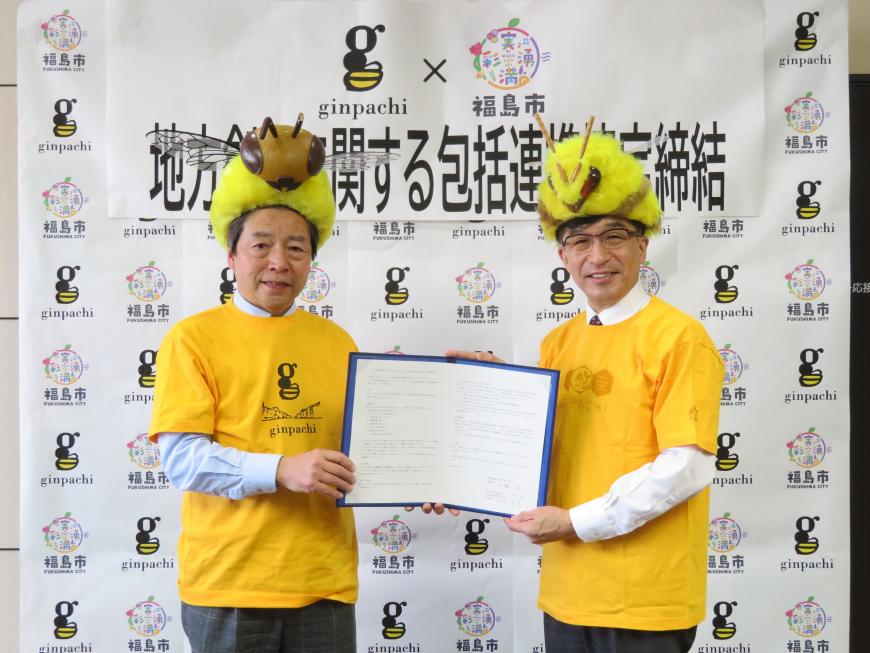 福島市とみつばちがデザインされたバックパネルの前で、みつばちの帽子を被り、黄色のTシャツを着た市長と男性で協定書を持っている写真