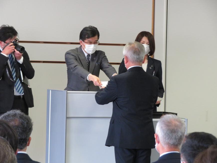 職員が座っている中、市長が男性に辞令を渡している写真
