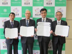 バックパネルの前で、市長を含む男性4名がそれぞれ協定書のホルダーをひろげて横一列に並んでいる写真