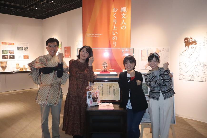 ガラスケースに入った土偶の前で古代の服を着た市長と菊池桃子さんと女性2名がそれぞれ腕をクロスさせたポーズをとっている写真