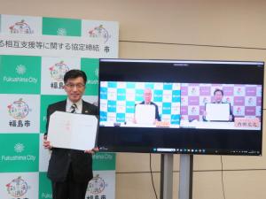 バックパネルの前に立つ市長と、モニターに映った2名の男性がそれぞれ協定書のホルダーをひろげている写真