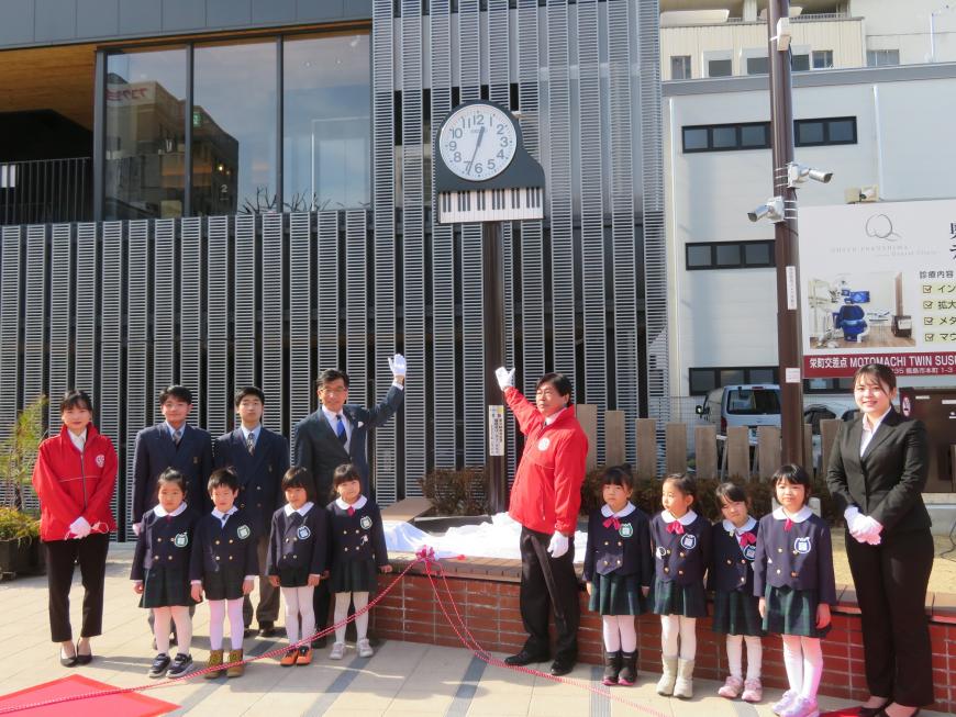 市長の他、参加した園児や関係者の方がピアノ型の特性時計の横に立ち、市長と男性が時計に手を向けている写真
