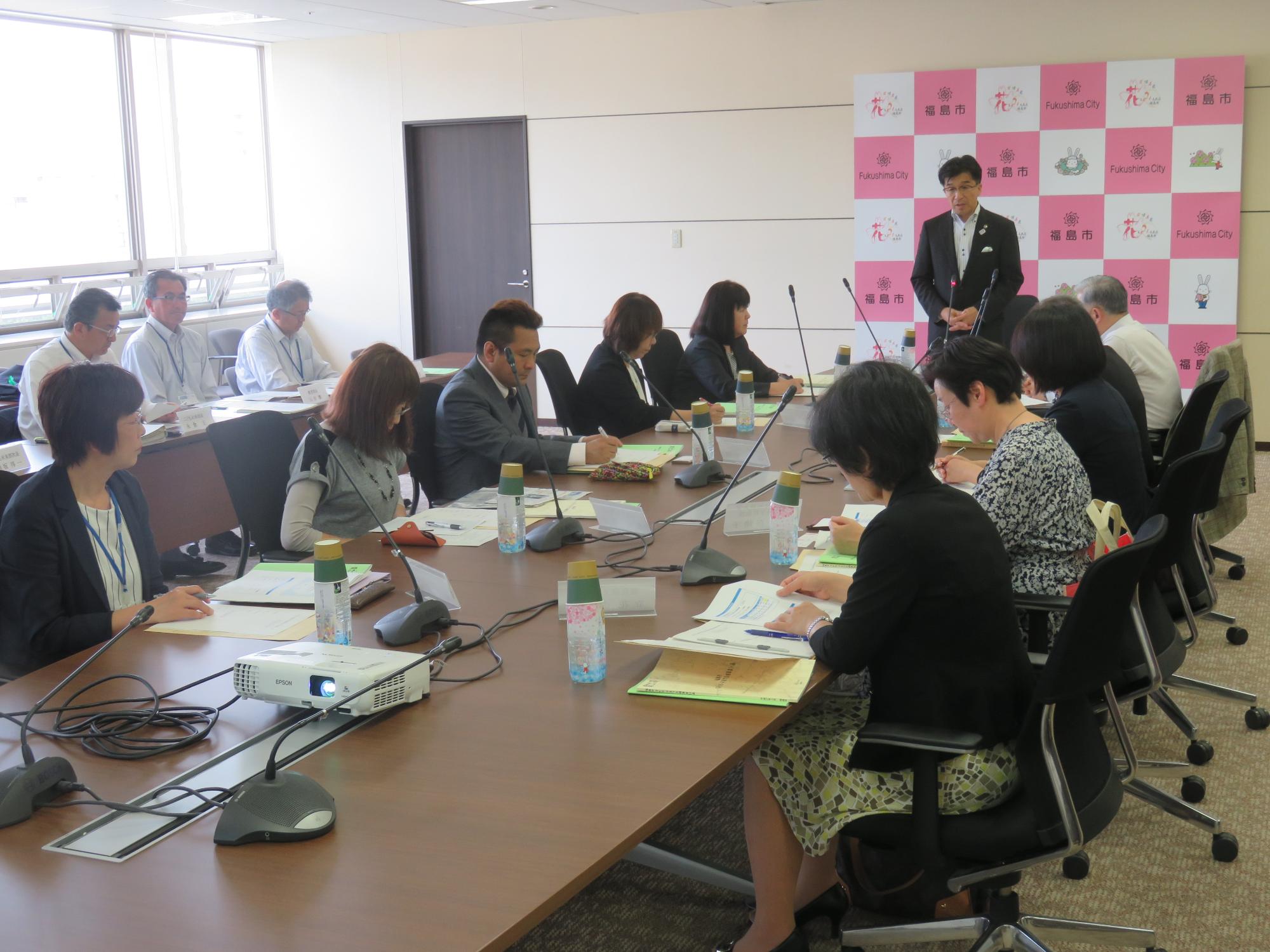 市役所庁議室の長机に関係者が向い合せで10数名椅子に座っており、正面奥に市長が立って話をしている写真