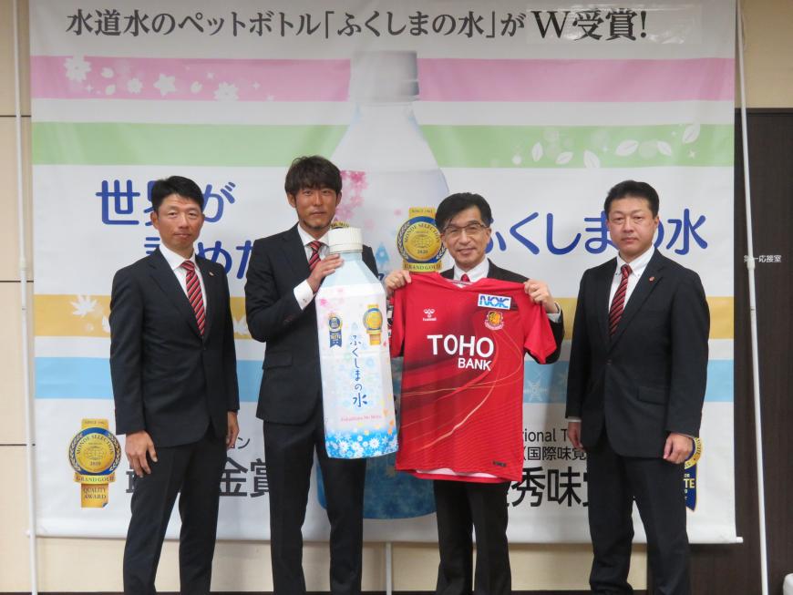 ふくしまの水の横断幕の前で市長が赤のユニフォームを広げ、男性がふくしまの水の巨大なペットボトルを持ち、両脇に男性が1名立っている写真