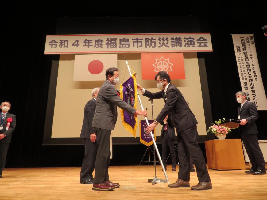 「福島市防災講演会」と書かれた横断幕があるステージで、市長が男性に旗を渡している写真