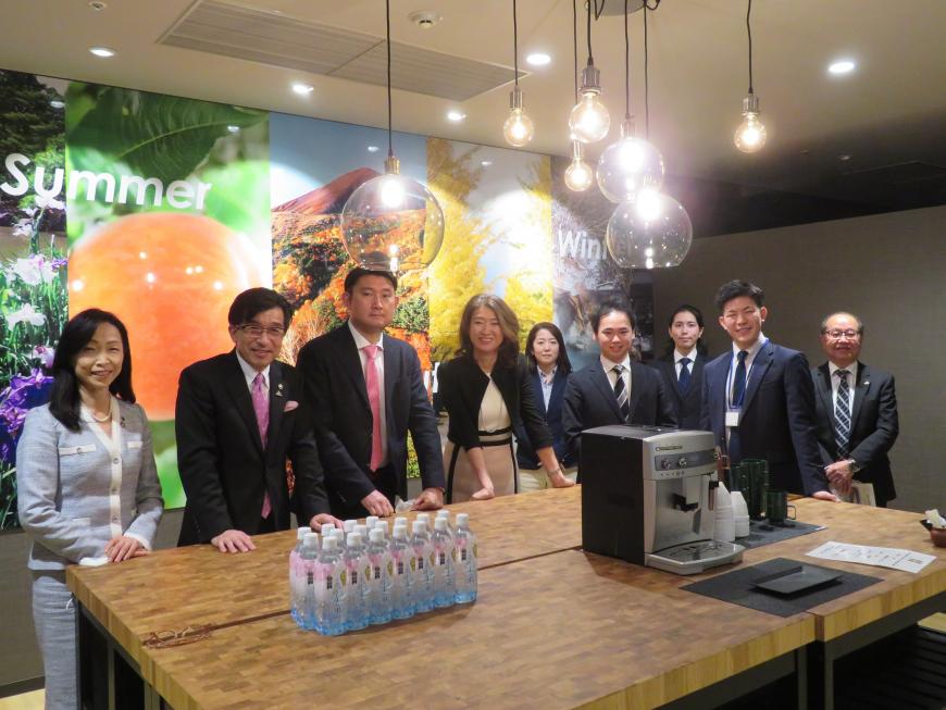 ペットボトルに入った飲料水や他の飲料を作る機械が置かれた机の回りに市長を含めて10人ほどが立ち撮影した写真