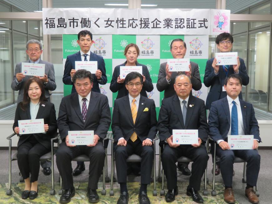 「福島市働く女性応援企業認証式」と書かれた横断幕が貼ってあるバックパネルの前で、認証書を持った男性7名、女性2名が市長を中心にしてひな壇状に2列に並んで撮影した写真