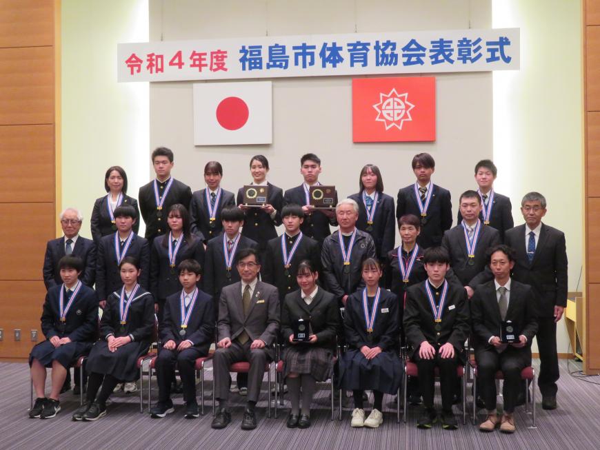 「福島市体育協会表彰式」と書かれた横断幕をバックにメダルを下げた男女、盾を持った男女合計20名程が最前列の方は市長を中心にして座り、ひな壇状に3列に並んで撮影した写真