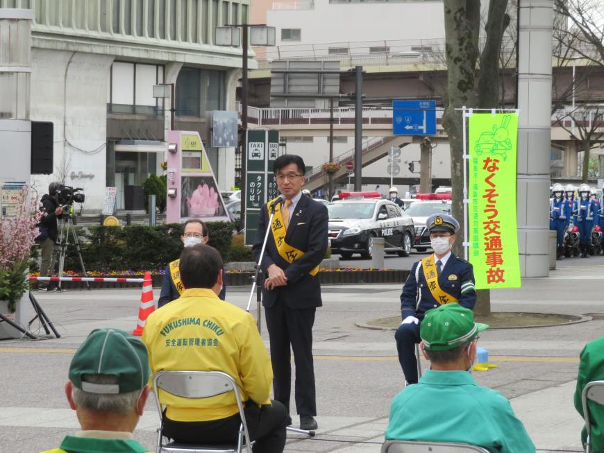 屋外の広場で交通安全の襷をかけた市長が座っている関係者の方々に話をしている写真