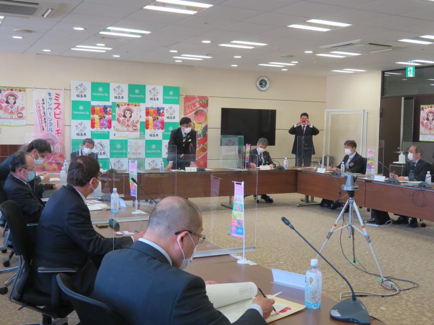 四角に並べられた席に出席者が座り、中央の位置の市長が自席で立ち話をしている写真