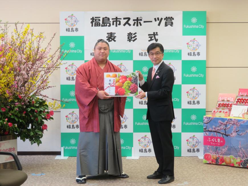 福島市スポーツ賞表彰式の横断幕が貼られたバックパネルの前で、市長と袴、羽織姿の若隆景関が果物のイラストのボードと目録封筒をを持っている写真