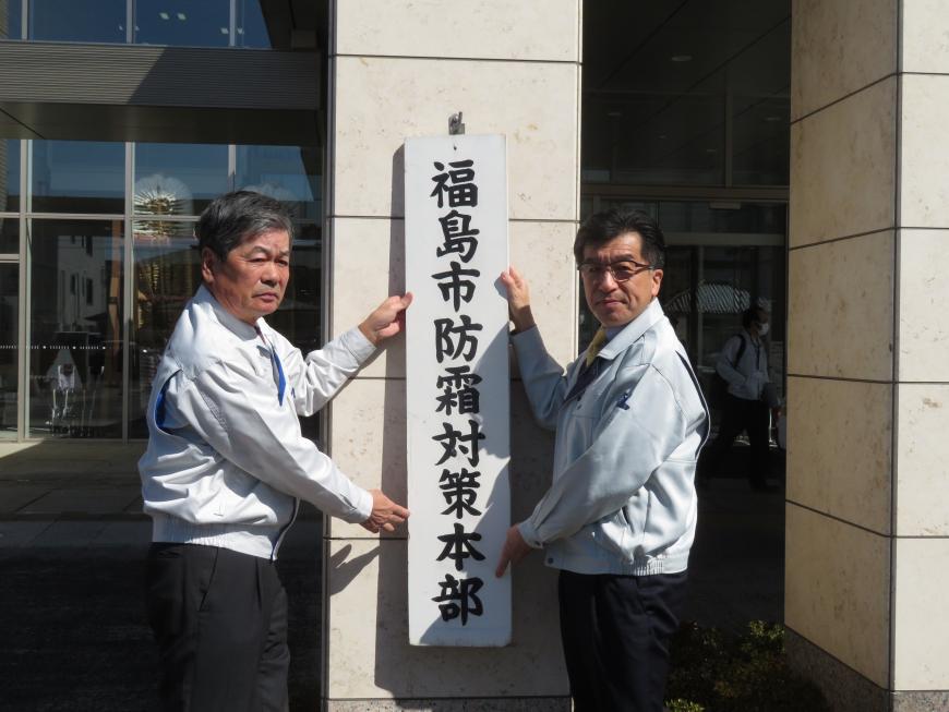 「福島市防霜対策本部」と書かれた看板を市長と男性で壁に掛けようとしている写真