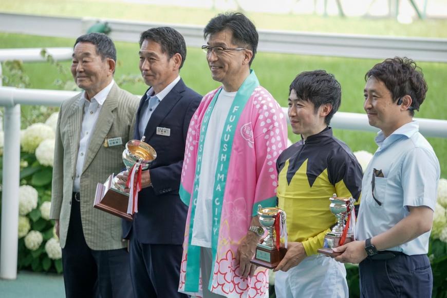 競馬場で市長の両脇に騎手を含めて男性2名ずつが立ち、その内3名はトロフィーを持っている写真