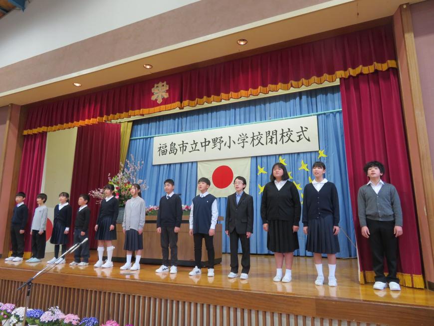 福島市立中野小学校閉校式と書かれた横断幕が吊るされたステージに、在校生が横一列に並んでいる写真