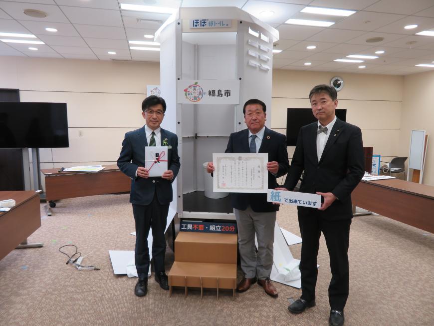 備蓄型組立式個室トイレの前で目録の封筒を持った市長と感謝状を持った男性と紙で出来ていますと書かれたプレートを持った男性が並び記念撮影している写真