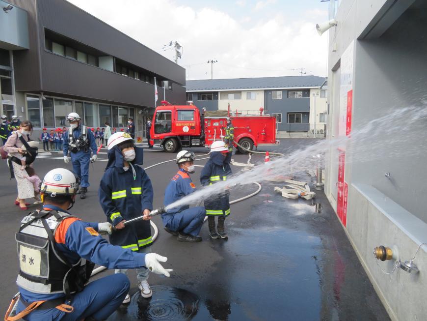 消防団員が補助しながら防火服を着た園児が放水訓練をしている写真