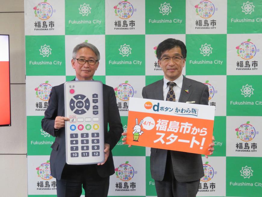 バックパネルの前で、市長が「福島市からスタート」と書かれたボードを持ち、男性が大きなリモコンのレプリカを持って「d」を指さしている写真