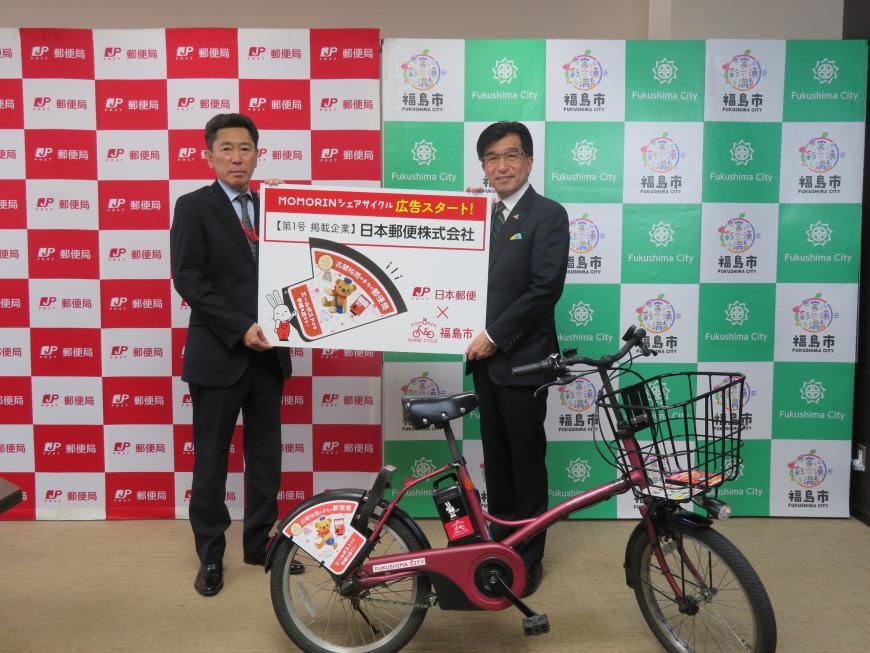 福島市と郵便局のバックパネルの前で、市長と男性で広告スタートを知らせるボードを持ち、その前に広告をつけた自転車を置いている写真