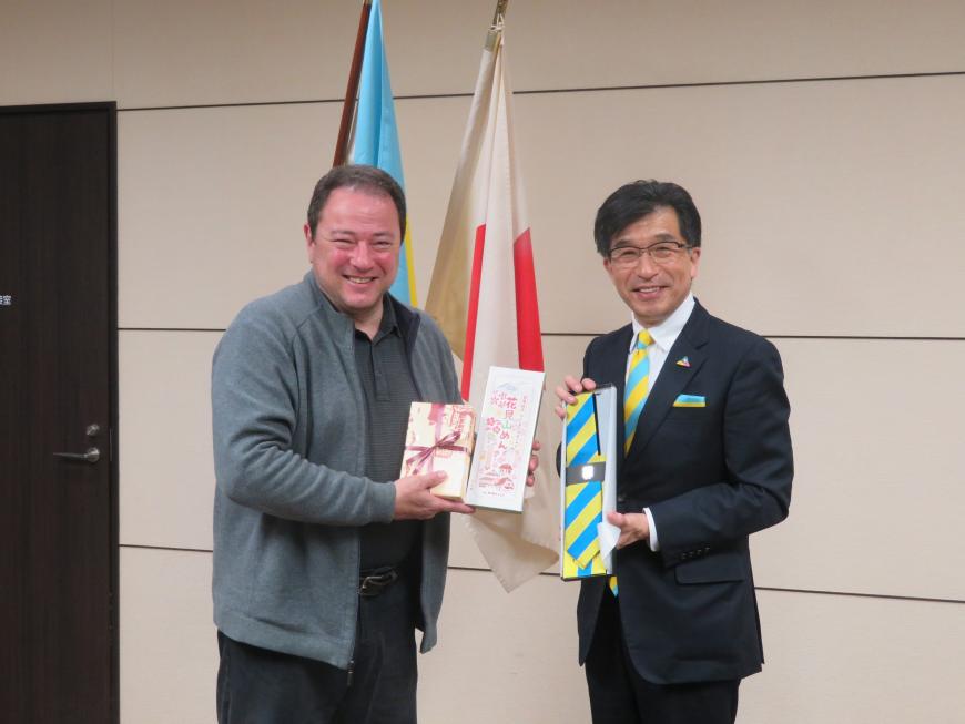 駐日大使が市からのお土産品を持ち、市長がウクライナ国旗の色のストライプのネクタイを持って立っている写真