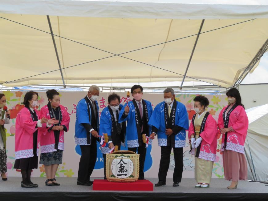テント内のステージで、青の法被を着た市長と男性3人、その両脇にピンクの法被を着た女性2人が立ち、男性4人で1つの酒樽を鏡開きをしている写真