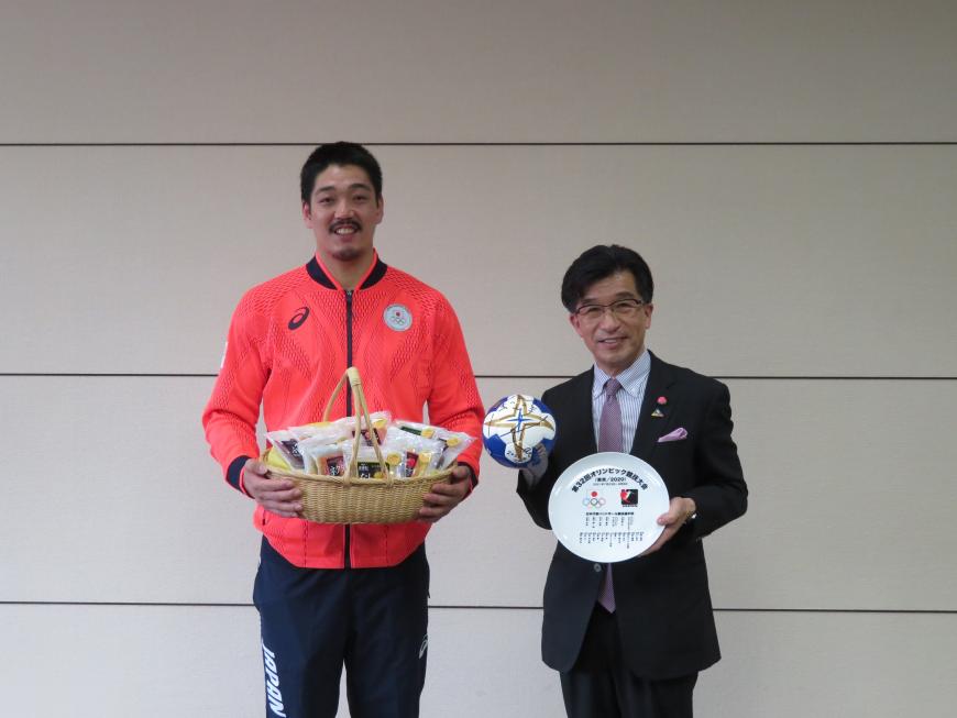 市長が左手に記念の皿、右手にハンドボールを持ち、日本代表のジャージ姿の男性が食品が入ったかごを持って並んでいる写真